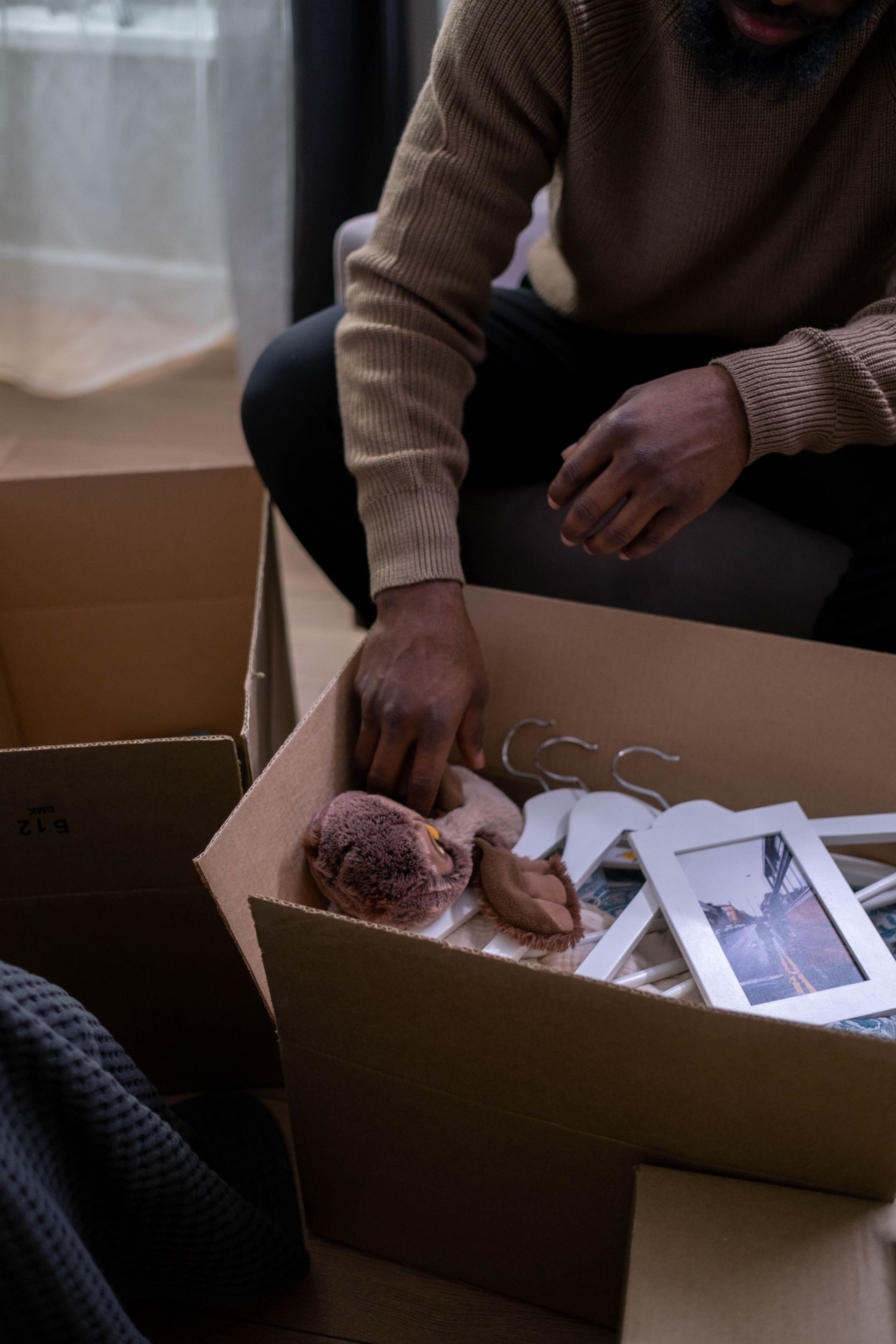 African adult unpacking personal belongings, including plush toy and pictures, in a cardboard box indoors.